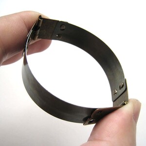 Könnte beinhalten: Ein ovales Metallarmband im Vintage-Stil mit einem Scharnierverschluss. Das Armband hat eine dunkle, antike Bronzefarbe und scheint aus dünnem, flexiblem Metall gefertigt zu sein. Der Verschluss wird mit kleinen Schrauben befestigt.