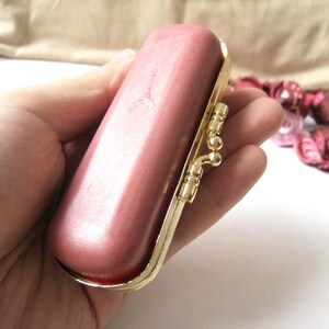 Può includere: Una pochette rettangolare rosa vintage con una cornice e chiusura color oro. La chiusura ha un design decorativo e ornato. La pochette è tenuta in mano da una persona, con uno sfondo neutro.