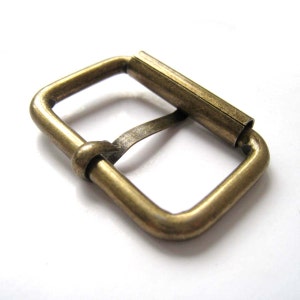 Pode incluir: Fivela metálica retangular com acabamento bronze. Possui uma barra de rolo e uma lingueta para prender as correias. O design é simples e funcional, adequado para vários projetos de artesanato e marroquinaria.