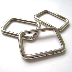 May include: Three shiny, silver metal rectangular rings. The rings are made of a smooth, reflective material and are arranged in a slightly overlapping pattern against a white background. The rings have rounded corners and a small seam.