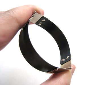 Op de afbeelding: Een zwart metalen band met een zilveren sluiting. De band is flexibel en kan worden aangepast aan verschillende polsmaten.