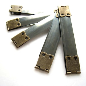 Op de afbeelding: Vijf metalen banden met messing gespen. De banden zijn zilverkleurig en hebben een licht getextureerd oppervlak. De gespen zijn glanzend messingkleurig en hebben een decoratief ontwerp.