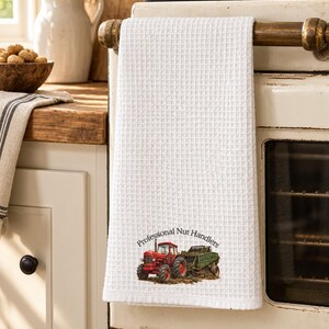 May include: A white waffle-weave kitchen towel featuring a graphic of a red tractor pulling a green harvester. The text "Professional Nut Handlers" is printed in a black arc above the tractor illustration on the lower portion of the towel.
