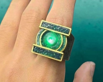 May include: A metallic ring featuring a rectangular gold-toned frame with dark green textured inlays. The center of the ring displays a circular, glowing green light element that mimics a lantern symbol, set against a dark, weathered metal band.