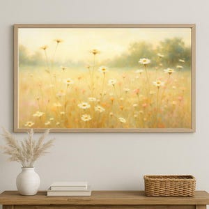 Puede incluir: Una obra de arte enmarcada representa un campo de margaritas blancas bajo un cielo amarillo suave. La pintura tiene una paleta de colores cálidos y un marco de madera marrón claro. La obra de arte se exhibe sobre una mesa de madera con un jarrón, libros y una cesta tejida.