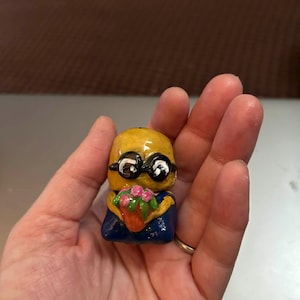 Op de afbeelding: Een kleine, handgeschilderde figuur van een Minion-personage met een boeket roze bloemen. Het beeldje heeft een gele body, een zwarte bril en een blauwe outfit. De bloemen zitten in een bruine pot.
