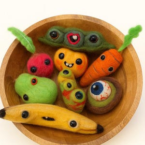 May include: A wooden bowl containing quirky felted vegetables, each with large, black button eyes. The collection includes a green pea pod, orange carrot, yellow banana, and other colourful, whimsical produce.