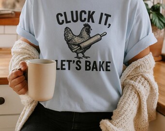 May include: Light blue t-shirt with the text "CLUCK IT, LET'S BAKE" and a graphic of a chicken holding a rolling pin. The shirt is worn with black shorts and a cream-colored cardigan. A beige mug is held in the hand.