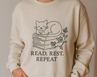 May include: Cream-colored sweatshirt featuring a black line drawing of a sleeping cat atop a stack of books, with ivy accents. The text "READ. REST. REPEAT" is printed below the illustration. The sweatshirt has long sleeves and a crew neck.