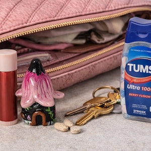 May include: A pink quilted purse with a gold zipper, a red lip balm, a decorative flower-shaped figurine, a bottle of Tums Ultra 1000 Berry Fusion, and a set of keys. Several antacid tablets are scattered nearby.