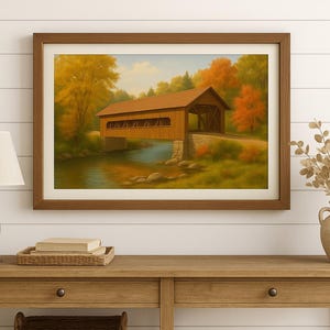 Puede incluir: Pintura enmarcada de un puente cubierto sobre un río, rodeado de árboles con follaje otoñal. La pintura está en un marco de madera marrón, expuesta sobre una mesa de madera.