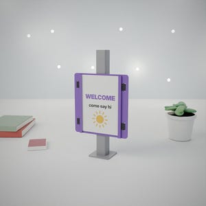 May include: A purple sign holder on a gray stand displays a white sign with the text WELCOME come say hi and a yellow sun icon. The sign is held by black clips on the sides. The display is positioned between stacked books and a potted plant.
