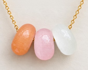 May include: A gold chain necklace featuring three smooth, oval-shaped cat's eye beads in orange, light pink, and white. The beads are strung horizontally along the center of the delicate gold metal chain, creating a simple and colorful pendant design.