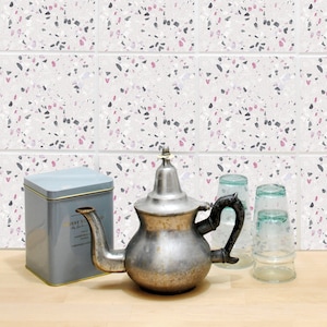 Puede incluir: Una tetera de plata con asa y pico, una lata azul con la palabra "NECTAR" en ella, y dos vasos de vidrio transparentes sobre una superficie de madera marrón claro. El fondo es una pared de azulejos blancos con un patrón de terrazo rosa y gris.