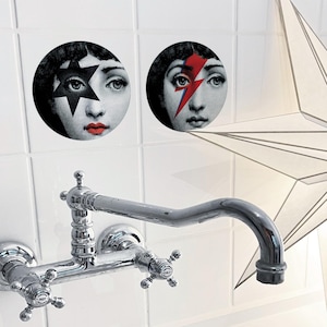 Puede incluir: Dos baldosas decorativas redondas con la cara de una mujer con un diseño de estrella y rayo. Un grifo cromado está montado en una pared de azulejos blancos. Una gran lámpara en forma de estrella blanca en el fondo.