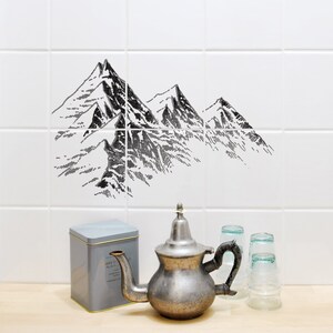 Peut inclure: Un mur composé de six carreaux blancs carrés arborant une illustration de chaîne de montagnes en noir et blanc. Sous les carreaux se trouvent une théière en métal argenté, une boîte de rangement grise et deux verres sur une surface en bois.