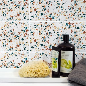 Puede incluir: Azulejos de cerámica blanca con un patrón de terrazo en tonos marrones, verde azulado y blanco. Una bandeja rectangular blanca con una esponja vegetal marrón, un tubo de crema de manos etiquetado como "Ahava" y una botella de gel de ducha etiquetada como "Ahava Mineral Botanic" están en la bandeja. Una toallita gris está en el lado derecho de la bandeja.