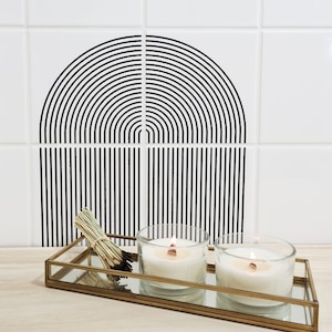 Puede incluir: Dos velas blancas en tarros de vidrio están sentadas en una bandeja dorada con fondo de espejo. La bandeja está sobre una superficie de madera clara. Un manojo de ramitas secas está en la bandeja. Un diseño geométrico en blanco y negro está en la pared detrás de la bandeja.