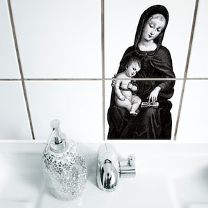 Puede incluir: Una imagen en blanco y negro de una mujer sosteniendo a un niño. La mujer lleva una túnica larga y el niño está sentado en su regazo. La imagen está impresa en azulejos blancos en un baño. Un dispensador de jabón plateado y un grifo plateado están en primer plano.