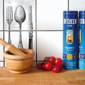 Puede incluir: Dos latas azules de espaguetis De Cecco, una con el texto "Spaghetti - 12 Cottura 12 min" y la otra con "ti - 12", sobre una encimera de madera con un mortero y un mazo de madera, un cuenco de tomates y un salpicadero de azulejos blancos con una ilustración en blanco y negro de un tenedor, una cuchara y un cuchillo.