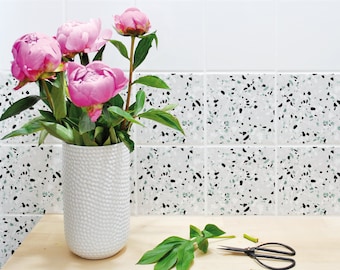 May include: A white ceramic vase with a textured surface holds a bouquet of pink peonies. The vase is sitting on a wooden table in front of a white tiled wall with a terrazzo pattern.