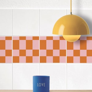 Puede incluir: Un salpicadero de cocina de azulejos blancos con un patrón de tablero de ajedrez en naranja y rosa. Una lámpara colgante amarilla cuelga sobre una taza de cerámica azul con la palabra "LOVE" impresa en ella.