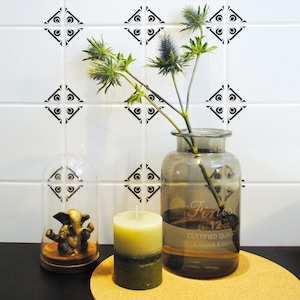 Puede incluir: Un jarrón de vidrio gris con una planta verde y una vela verde y blanca sobre un posavasos de corcho. El jarrón tiene el texto "Finest Fl. 12 oz. Certified Quality Limited Edition". El jarrón está delante de una pared de azulejos blancos con diseños geométricos negros.