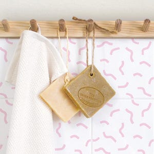 May include: White bathroom tiles with a pink abstract pattern of wavy lines. Two bars of soap hang from hooks on a wooden rack above the tiles.