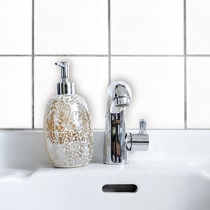 Puede incluir: Un lavabo de baño de cerámica blanca con un grifo cromado y un dispensador de jabón de vidrio decorativo. El dispensador de jabón es un patrón de mosaico con acentos dorados y blancos.
