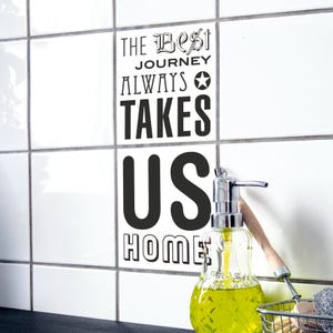 Könnte beinhalten: Ein weißer Fliesenaufkleber mit dem schwarzen Text THE BEST JOURNEY ALWAYS TAKES US HOME und einem Sternsymbol. Der Aufkleber ist auf weißen quadratischen Fliesen neben einem durchsichtigen Seifenspender mit gelber Flüssigseife angebracht.