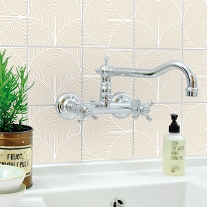 Puede incluir: Un lavabo de baño blanco con un grifo cromado y un salpicadero de azulejos blancos con un patrón geométrico en dorado. Una botella de jabón de manos está en la encimera con el texto "Detenga el agua mientras me usa".