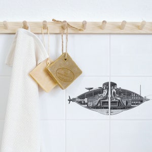 May include: A wooden rack with hooks holds a white towel, two bars of soap, and a string of twine. The soap bars are square and have a brown colour. The soap bar on the right has the text "The North Star Soap Co. Australia" printed on it. The background is white tile with a black and white illustration of a submarine.