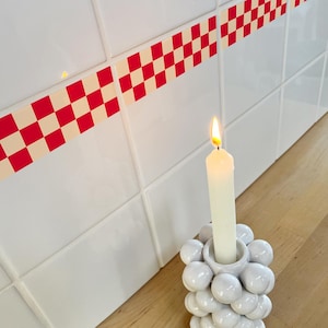 Può includere: Una candela bianca accesa in un portacandele in ceramica bianca con un design a bolle. Il portacandele è appoggiato su una superficie di legno accanto a una parete piastrellata bianca con un bordo a scacchi rosso e bianco.