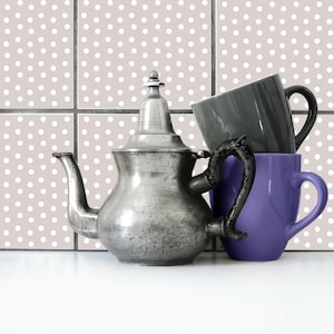 Puede incluir: Una tetera de plata con asa negra y una taza morada a su lado. Ambas están sobre una superficie de azulejos blancos con un patrón de lunares.