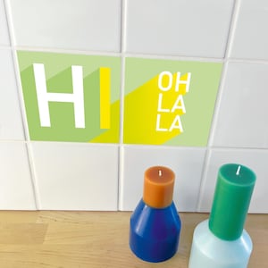 Puede incluir: Dos velas decorativas, una azul y naranja, la otra verde y blanca. Arriba, dos baldosas cuadradas muestran las palabras "HI" y "OH LA LA" en blanco y amarillo sobre un fondo verde. Las velas están sobre una superficie de madera.