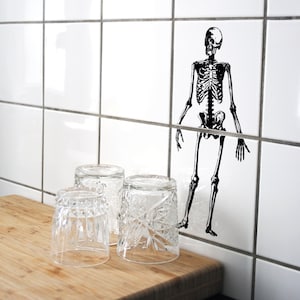 Puede incluir: Tres vasos de vidrio transparentes con un patrón decorativo se encuentran sobre una superficie de madera. Un adhesivo de esqueleto en blanco y negro está en la pared de azulejos blancos detrás de los vasos.