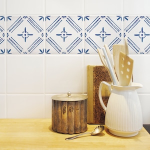 Può includere: Un'anfora di ceramica bianca con un designo a scanalature si trova su un bancone di legno. Un contenitore di metallo argentato con un coperchio si trova a sinistra dell'anfora. Un libro e utensili da cucina in legno si trovano dietro l'anfora. Lo sfondo è una parete piastrellata bianca con un bordo a motivi blu e bianchi.