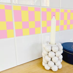 Puede incluir: Un portavelas de cerámica blanca con un grupo de formas redondas en forma de burbujas. Una vela alta y blanca está insertada en la parte superior. El fondo presenta un patrón de cuadros rosas y amarillos y azulejos blancos.