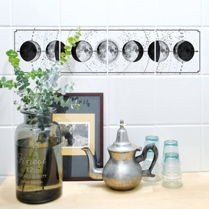 Puede incluir: Un adhesivo de fases lunares en blanco y negro en una pared de azulejos blancos. El adhesivo presenta ocho fases de la luna, conectadas por una línea punteada. El adhesivo está sobre una tetera plateada, un jarrón con vegetación y dos vasos de vidrio transparentes.