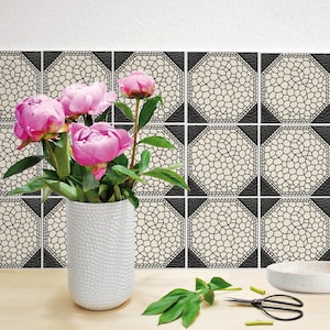 Puede incluir: Un jarrón blanco texturizado contiene peonías rosas con hojas verdes. Detrás del jarrón hay baldosas decorativas con un patrón de mosaico negro y crema. Un cuenco pequeño y unas tijeras están sobre la superficie de madera.