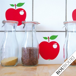 Puede incluir: Tres botellas de vidrio transparente con tapas, llenas de diferentes granos, sobre una encimera de madera. Las botellas están delante de una pared de azulejos blancos con pegatinas de manzanas rojas.