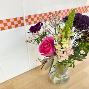 May include: A bouquet of pink, white, and purple flowers in a clear glass vase with a textured surface. The flowers are arranged on a wooden countertop with a pink and orange checkered tile border.