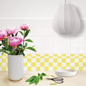 Puede incluir: Un jarrón de cerámica blanco con una superficie texturizada lleno de peonías rosas. El jarrón está sobre una encimera con un salpicadero de azulejos en forma de tablero de ajedrez amarillo y blanco. También hay un cuenco blanco y unas tijeras de jardín en la encimera.