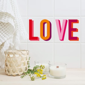 Puede incluir: Decoración mural con la palabra "LOVE" en letras 3D rojas, naranjas, rosas y magenta. La escena incluye una cesta tejida, dos velas en portavelas de cristal y una ramita de flores amarillas. Una toalla blanca cuelga en el fondo.