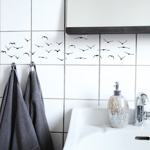 Puede incluir: Azulejos de baño blancos con siluetas de pájaros negros, dos toallas grises colgadas de ganchos, un lavabo blanco, un dispensador de jabón dorado y un grifo cromado.