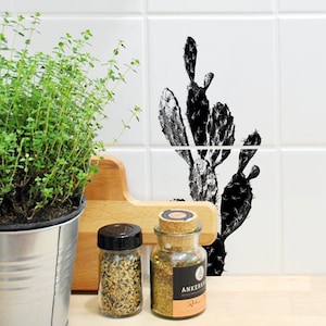 Op de afbeelding: Zwart-witte cactus illustratie op witte tegels met twee glazen potten gevuld met kruiden en een plant in een pot op de voorgrond.