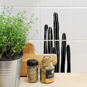Puede incluir: Una encimera de cocina de azulejos blancos con una planta en maceta, una tabla de cortar de madera y dos frascos de vidrio con tapas de corcho. Los frascos contienen especias y están etiquetados como "Ankerkraut" y "Rubin Pfeffer". Un diseño de cactus en blanco y negro está en la pared de azulejos detrás de los frascos.