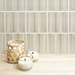 Può includere: Tre candele in vasetti di vetro, due con stoppini di legno e una in un cesto intrecciato, su una superficie di legno chiaro davanti a un muro di piastrelle rettangolari beige.
