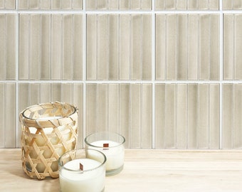 May include: Three candles in glass jars, two with wooden wicks, and one in a woven basket, on a light wood surface in front of a wall of beige rectangular tiles.