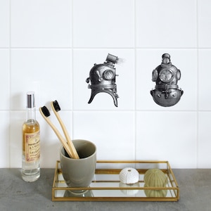 Puede incluir: Dos ilustraciones en blanco y negro de cascos de buceo vintage en una pared de azulejos blancos. Los cascos están en un mostrador de baño con una botella de vidrio transparente, dos cepillos de dientes de madera, una taza de cerámica gris y dos erizos de mar en una bandeja dorada.